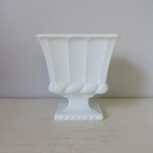 May include: A white, vintage-style urn with a square base and a decorative rope-like design. The urn has a fluted body and a flared top. The piece is made of a solid, opaque material and sits on a white surface.