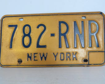 May include: Vintage New York license plate with the number 782-RNR in dark blue against a yellow background. The word "NEW YORK" is printed in blue at the bottom. The plate has four holes and a rectangular box.