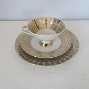 May include: A white teacup and saucer set with gold geometric patterns. The cup has a wide, shallow bowl and a delicate handle. The saucer has a matching pattern and a slightly raised rim.