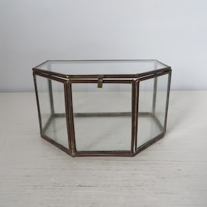 May include: A vintage, geometric glass display box with a hinged lid. The box has a brass-colored metal frame and is composed of multiple glass panels. The box is octagonal in shape and sits on a white surface.