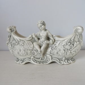 May include: A white ceramic planter shaped like a boat with a cherub figure sitting in the center. The planter has intricate floral designs and a gray glaze.