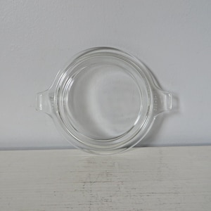 May include: A clear glass Pyrex lid with two small handles. The lid is round with concentric rings and is designed to fit a Pyrex dish. The glass is transparent, allowing visibility of the contents.