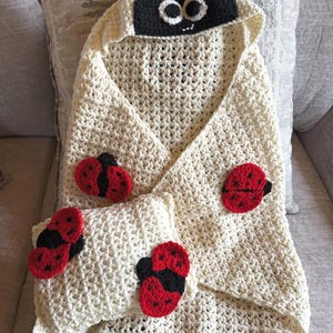 Op de afbeelding: Een crèmekleurige, gehaakte babydeken en kussenset. De deken heeft een capuchon met een zwart lieveheersbeestjesgezicht en een rode strik. Rode en zwarte lieveheersbeestjes applicaties zijn op de deken en het kussen genaaid.