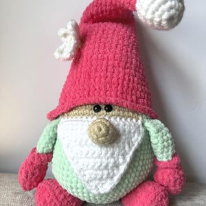 Könnte beinhalten: Eine handgefertigte gehäkelte Zwergpuppe mit rosa Hut und passenden Fäustlingen und Füßen. Der Zwerg hat einen hellgrünen Körper, einen weißen Bart und eine beige Nase. Der Hut hat eine weiße Bommel und eine weiße Blumenverzierung.