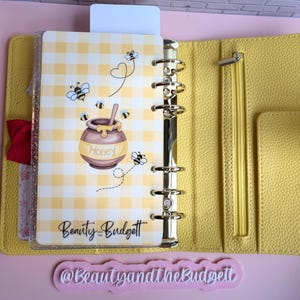 May include: A yellow textured six-ring binder featuring a clear dashboard with a yellow gingham pattern, cartoon bees, and a honey pot illustration. The text "Honey" and "Beauty and the Budget" are printed on the dashboard. A zipper pocket is visible.
