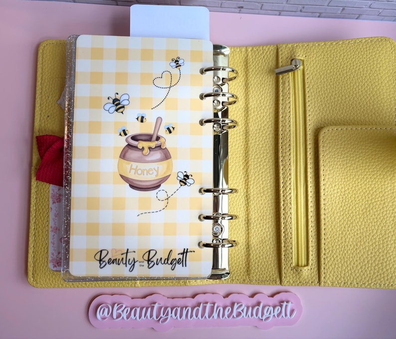 May include: A yellow textured six-ring binder featuring a clear dashboard with a yellow gingham pattern, cartoon bees, and a honey pot illustration. The text "Honey" and "Beauty and the Budget" are printed on the dashboard. A zipper pocket is visible.