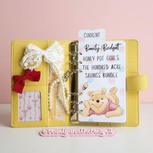 May include: A yellow six-ring binder featuring a white lace bow, a small red bow, and a pearl charm. The insert displays the text: CURRENT Beauty and the Budget HONEY POT GOALS: THE HUNDRED ACRE SAVINGS BUNDLE, with a cartoon bear illustration.