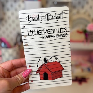 May include: A white, lined, six-hole punched planner insert featuring the text "Beauty and the Budget" at the top, followed by "Little Peanuts Savings Bundle" in black font, and a graphic of a white dog lying on a red doghouse at the bottom.