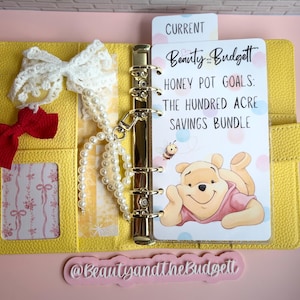 May include: A yellow six-ring binder featuring a white insert with the text Beauty and the Budget, Honey Pot Goals: The Hundred Acre Savings Bundle, and an illustration of a bear. Includes a white lace bow, a red bow, and a string of white pearls.