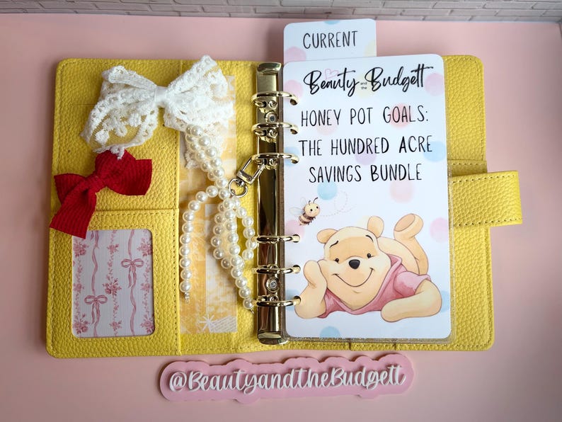 May include: A yellow six-ring binder featuring a white insert with the text Beauty and the Budget, Honey Pot Goals: The Hundred Acre Savings Bundle, and an illustration of a bear. Includes a white lace bow, a red bow, and a string of white pearls.