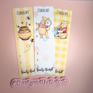 May include: Three vertical planner bookmarks featuring Winnie the Pooh illustrations. The left shows a honey pot, the center shows Pooh with a magnifying glass, and the right shows Pooh eating honey. Each has the word CURRENT and Beauty and the Budgett.