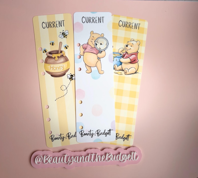 May include: Three vertical planner bookmarks featuring Winnie the Pooh illustrations. The left shows a honey pot, the center shows Pooh with a magnifying glass, and the right shows Pooh eating honey. Each has the word CURRENT and Beauty and the Budgett.