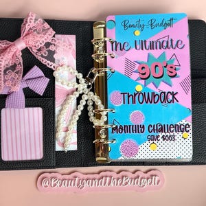 May include: A black leather-textured binder with gold rings, featuring a pink and blue insert titled The Ultimate 90's Throwback Monthly Challenge save $663. Includes a pink lace bow, a purple bow, a pearl bead charm, and a pink striped card.