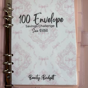 Könnte beinhalten: Ein hellrosa Ringbuch mit einer 100-Umschläge-Spar-Challenge-Einlage mit hellrosa Schleifenmuster. Der Text lautet 100 Envelope Savings Challenge Save $5050 und Beauty and the Budgett. Ein rosa Schild darunter zeigt @BeautyandtheBudgett.