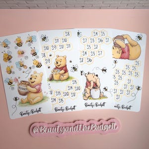 May include: Four white rectangular savings challenge sheets featuring Winnie the Pooh illustrations, honey pots, bees, and honeycomb patterns with dollar amounts. Each sheet includes the text Beauty and the Budget and a separate pink sign with the text @BeautyandtheBudget.