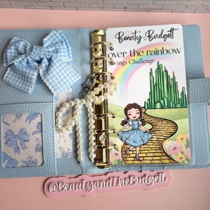 May include: A light blue textured binder featuring a gold six-ring mechanism, a blue gingham bow, and a pearl charm. Inside is a savings challenge card titled Beauty and the Budget Over the Rainbow, depicting a character on a yellow brick road.