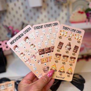 May include: Three rectangular sticker sheets with a light peach background featuring cartoon illustrations of marshmallows, chocolate, and campfires. The sheets are labelled Marshmallow Stacks, Stackin' S'more cash, and Toasty Little cash with various dollar amounts printed below each icon.