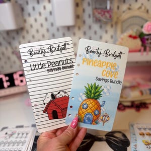 May include: Two rectangular paper savings trackers with punched holes. One features the text Beauty Budget, Little Peanuts Savings Bundle, and a cartoon dog on a red house. The other features Beauty Budget, Pineapple Cove Savings Bundle, and a pineapple house.