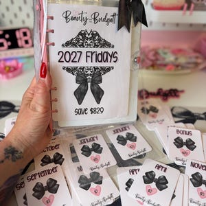 May include: A clear plastic binder with a black ribbon bow on the top right corner. The cover insert features the text Beauty and the Budget, 2027 Fridays, and save $829, decorated with black filigree and a bow graphic. Includes monthly cash envelopes.