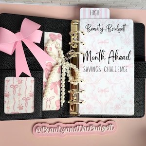 May include: A black textured binder with gold rings containing a Beauty and the Budget Month Ahead Savings Challenge insert. It features a pink bow, a white pearl beaded charm, and a pink and white floral ribbon pattern on the interior pockets.