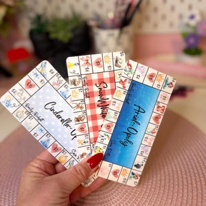 May include: Three rectangular paper savings trackers featuring themed designs labeled Cinderella, Snow White, and Ariel-Opoly. Each card displays a grid of icons with associated dollar amounts, including illustrations of crowns, hearts, and characters.