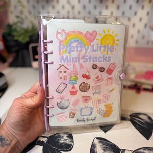 May include: A clear plastic binder with a light purple spine and snap closure. The front cover features a colourful illustration with the text "Pretty Little Mini Stacks" and "Beauty Budget" surrounded by icons including a rainbow, sun, heart, house, macarons, cupcake, coffee cup, television, and various other small decorative graphics.