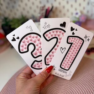 Peut inclure: Trois cartes rectangulaires blanches en papier présentant de grands contours noirs des chiffres 3, 2 et 1. Chaque chiffre est rempli de petites icônes de cœurs roses contenant des signes dollar. Les cartes incluent des cœurs et étoiles noirs, avec le texte save$44.