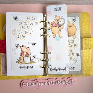 May include: A yellow six-ring binder containing white savings tracker inserts featuring Winnie the Pooh illustrations, honeycomb patterns with dollar amounts, and the text Beauty and the Budgett. A bookmark with the text CURRENT is included.