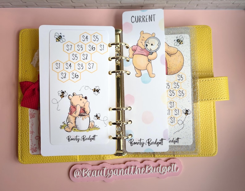 May include: A yellow six-ring binder containing white savings tracker inserts featuring Winnie the Pooh illustrations, honeycomb patterns with dollar amounts, and the text Beauty and the Budgett. A bookmark with the text CURRENT is included.
