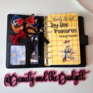 May include: A black leather-textured binder with gold rings containing a yellow savings bundle insert titled Toy Box Treasures featuring a cowboy illustration. Includes a multi-colored patterned bow, a small red bow, and a black beaded charm.