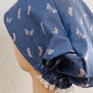 May include: A blue scrub cap featuring a pattern of light-colored moths and small white stars. The cap has a gathered back with white lace trim, a white ribbon tie, and a small heart-shaped charm attached to the end of the ribbon.
