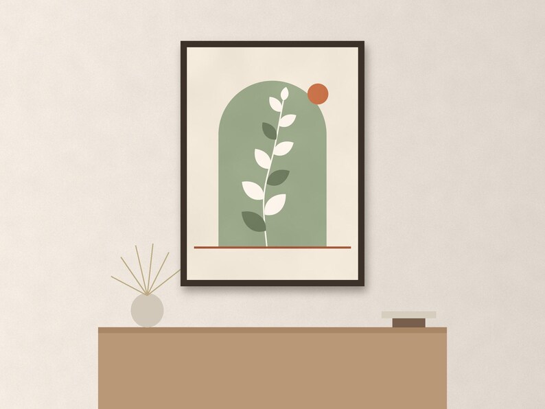May include: A framed wall art print featuring a sage green arch shape containing a white leafy plant stem. A small burnt orange circle sits in the upper right corner of the arch, and a thin horizontal red line runs across the bottom of the frame.