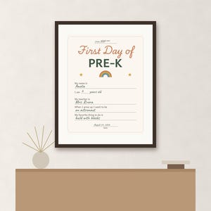 May include: A framed First Day of PRE-K sign with a cream background, dark brown frame, and rainbow graphic. The text includes fields for name, age, teacher, future career, and favorite activity, with handwritten details like Amelia and astronaut.
