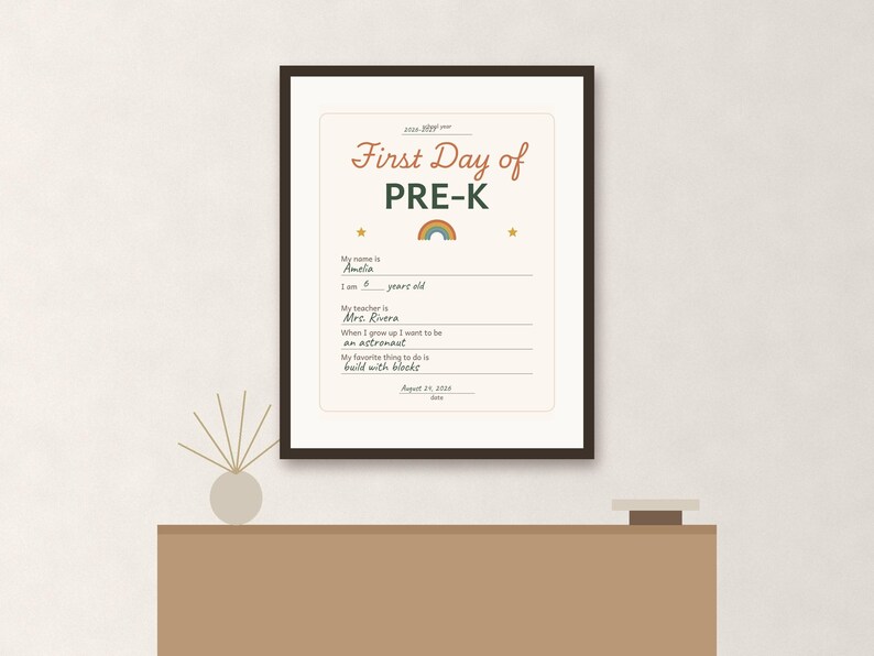 May include: A framed First Day of PRE-K sign with a cream background, dark brown frame, and rainbow graphic. The text includes fields for name, age, teacher, future career, and favorite activity, with handwritten details like Amelia and astronaut.