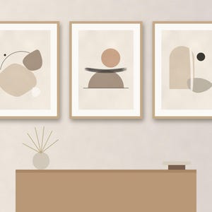 May include: Three framed wall art prints featuring abstract geometric shapes in shades of beige, tan, and brown. The prints are displayed in light wood frames. Below the art, a small round vase with thin sticks and a small square object sit on a table.