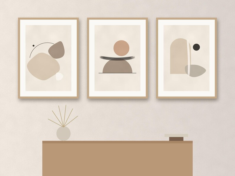 May include: Three framed wall art prints featuring abstract geometric shapes in shades of beige, tan, and brown. The prints are displayed in light wood frames. Below the art, a small round vase with thin sticks and a small square object sit on a table.