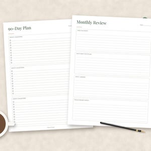 May include: Two white paper planning sheets featuring black text and lines. The left sheet is titled 90-Day Plan with sections for monthly milestones. The right sheet is titled Monthly Review with sections for monthly goals, lessons, and focus areas. A coffee cup and pen are present.