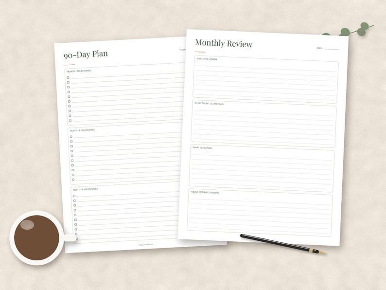 May include: Two white paper planning sheets featuring black text and lines. The left sheet is titled 90-Day Plan with sections for monthly milestones. The right sheet is titled Monthly Review with sections for monthly goals, lessons, and focus areas. A coffee cup and pen are present.