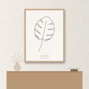 May include: A framed wall art print featuring a black line drawing of a monstera leaf on a cream background. The text MONSTERA and Monstera deliciosa is printed in black at the bottom. The frame is a light wood color with a thin profile.