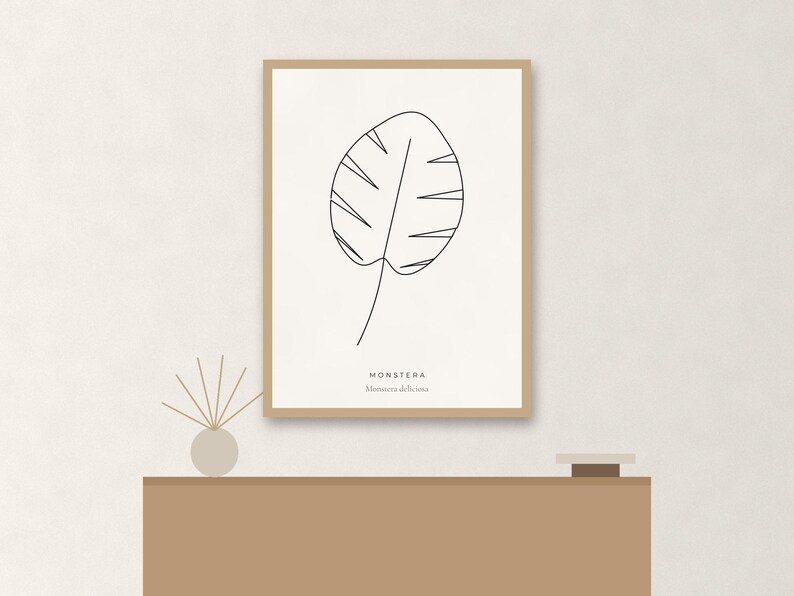 May include: A framed wall art print featuring a black line drawing of a monstera leaf on a cream background. The text MONSTERA and Monstera deliciosa is printed in black at the bottom. The frame is a light wood color with a thin profile.