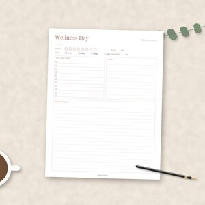 May include: A white Wellness Day planner sheet with sections for notes, mood, self-care, goals, and reflections. The page features a brown header, a cup of black coffee, and a black pen. A small green eucalyptus sprig is placed at the top right corner.
