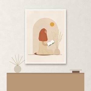 May include: A framed wall art print featuring a stylized illustration of a person wearing a rust-colored head covering and a beige garment, sitting and reading a book. The scene includes a yellow sun, an arched doorway, and a vase with dried grass.