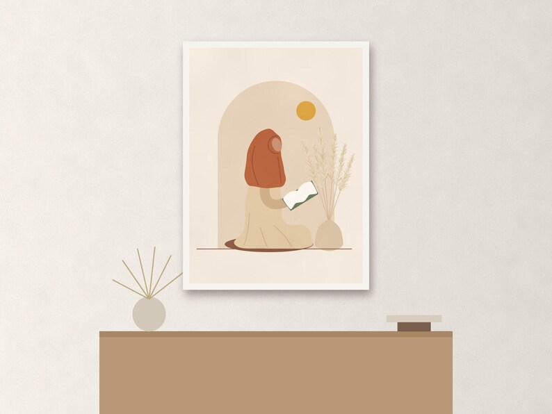 May include: A framed wall art print featuring a stylized illustration of a person wearing a rust-colored head covering and a beige garment, sitting and reading a book. The scene includes a yellow sun, an arched doorway, and a vase with dried grass.