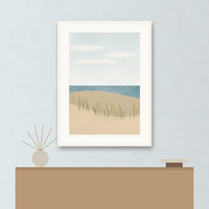 May include: A framed wall art print featuring a coastal landscape with a sandy beach, tall green dune grass, a blue ocean horizon, and a pale sky with soft white clouds. The artwork is displayed in a white frame, positioned above a brown console table.