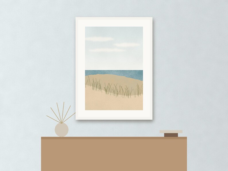 May include: A framed wall art print featuring a coastal landscape with a sandy beach, tall green dune grass, a blue ocean horizon, and a pale sky with soft white clouds. The artwork is displayed in a white frame, positioned above a brown console table.