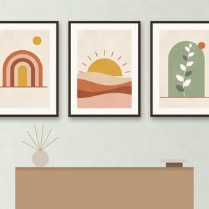 May include: Three framed wall art prints featuring abstract designs. The left print shows a rainbow and sun, the center print depicts a sun over layered hills, and the right print displays a green arch with a white leaf branch and a small red circle.