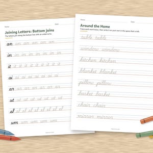 May include: Two white educational worksheets with black text for cursive handwriting practice. The left sheet is titled Joining Letters: Bottom joins, and the right sheet is titled Around the Home. Several colored crayons are placed around the papers.