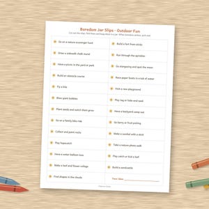 May include: A white rectangular sheet of paper titled Boredom Jar Slips - Outdoor Fun, featuring two columns of printed activity prompts with small sun icons, surrounded by several colorful wax crayons on a light brown wood-grain surface.