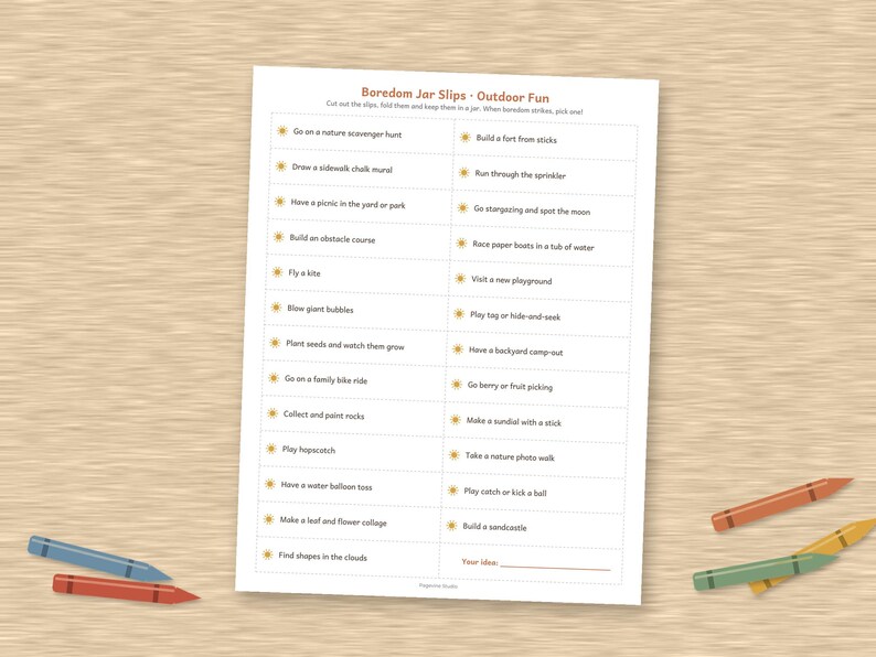 May include: A white rectangular sheet of paper titled Boredom Jar Slips - Outdoor Fun, featuring two columns of printed activity prompts with small sun icons, surrounded by several colorful wax crayons on a light brown wood-grain surface.