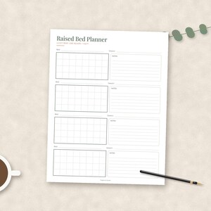 May include: A white paper sheet titled Raised Bed Planner with four sections, each containing a grid for garden layout and lined space for notes. The text reads 1 FOOT = 1 SQUARE / 1 GRID. A black pen and a small green leaf sprig are nearby.